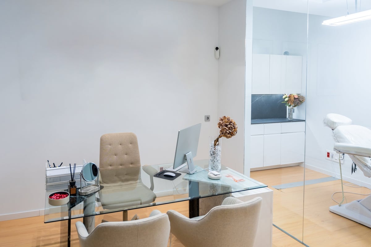 A modern, discreet aesthetic consulting room with a glass desk, ergonomic chairs and treatment bed in a calm neutral interior