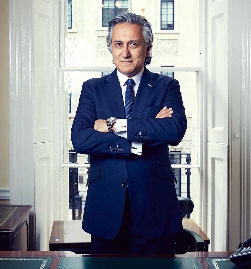 Middle-aged man in dark blue suit and tie standing with crossed arms in formal office setting with white doors and windows